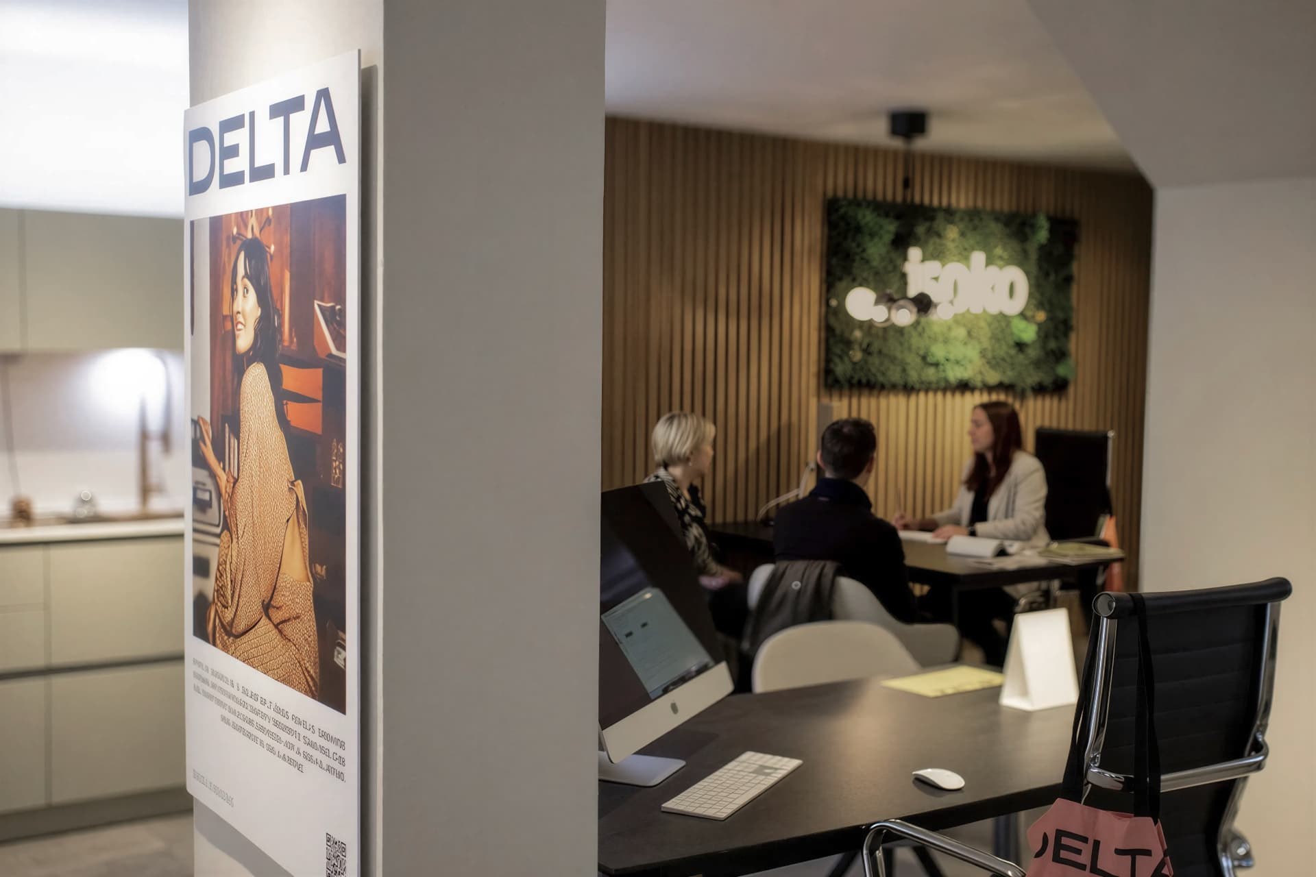 Sala de reuniones del estudio ISOKO: tres personas conversan alrededor de la mesa junto al panel de listones de madera; en primer término, la cocina de exposición