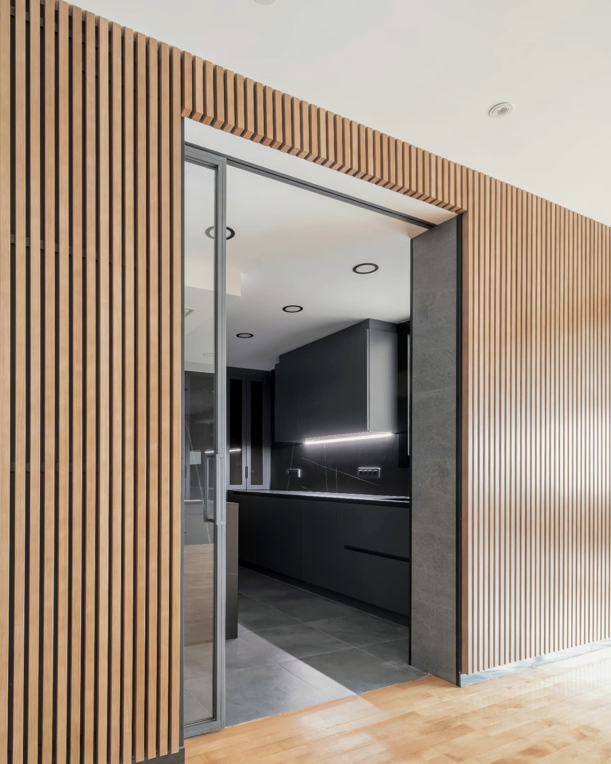 Paño continuo de listones de madera con hueco practicable hacia una cocina de frentes oscuros