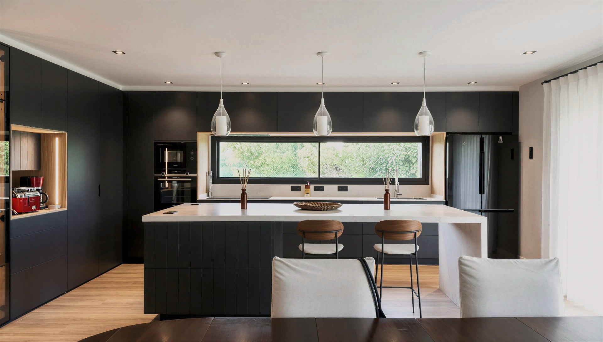 Cocina con isla negra, encimera clara, taburetes de madera y tres lámparas colgantes ante una ventana horizontal
