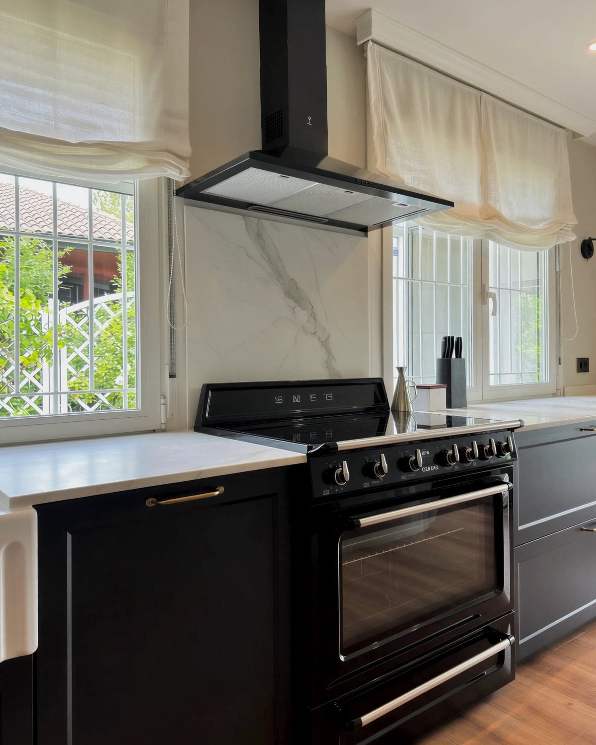 Cocina de gas negra bajo campana con frente de piedra clara veteada entre dos ventanas